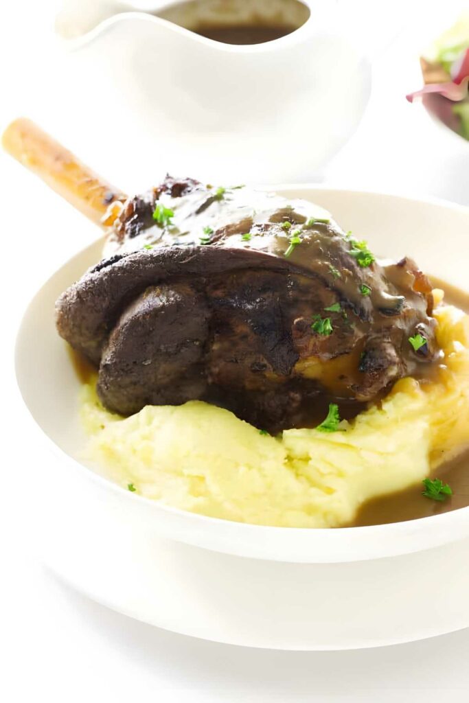 Lamb shank with gravy on mashed potatoes, garnished with parsley.