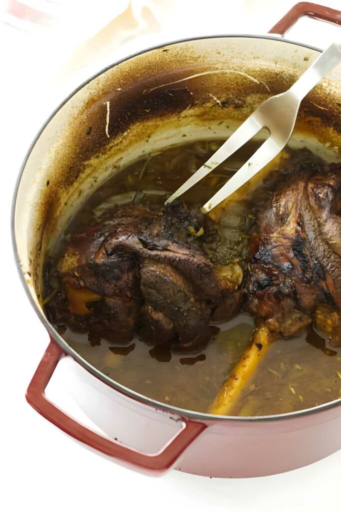 Braised lamb shanks in Dutch oven with broth, fork resting beside.