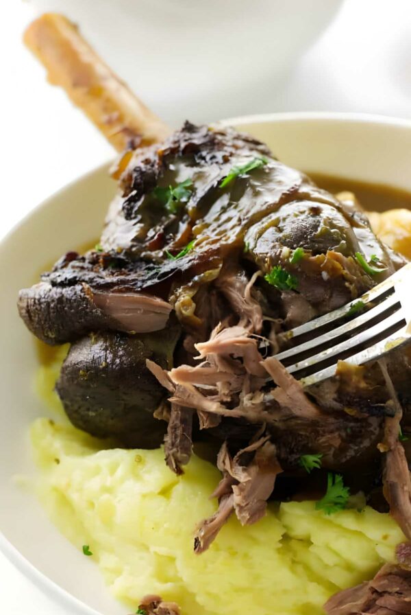 Braised lamb shank with mashed potatoes, gravy, parsley; fork pulling meat.