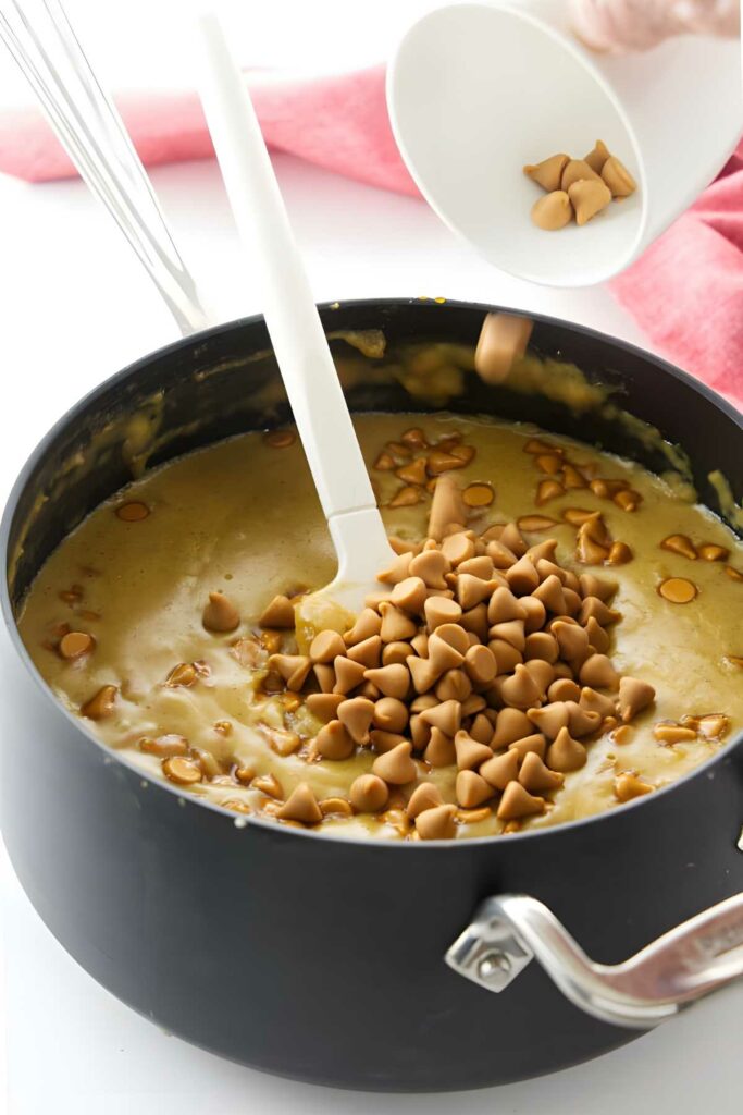 Hand pours butterscotch chips into saucepan of yellow batter with white spatula.
