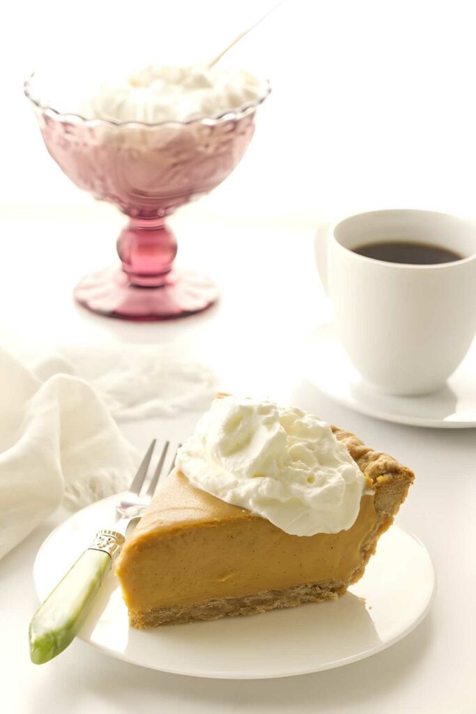 Pumpkin pie slice with whipped cream on plate, coffee and extra cream nearby.