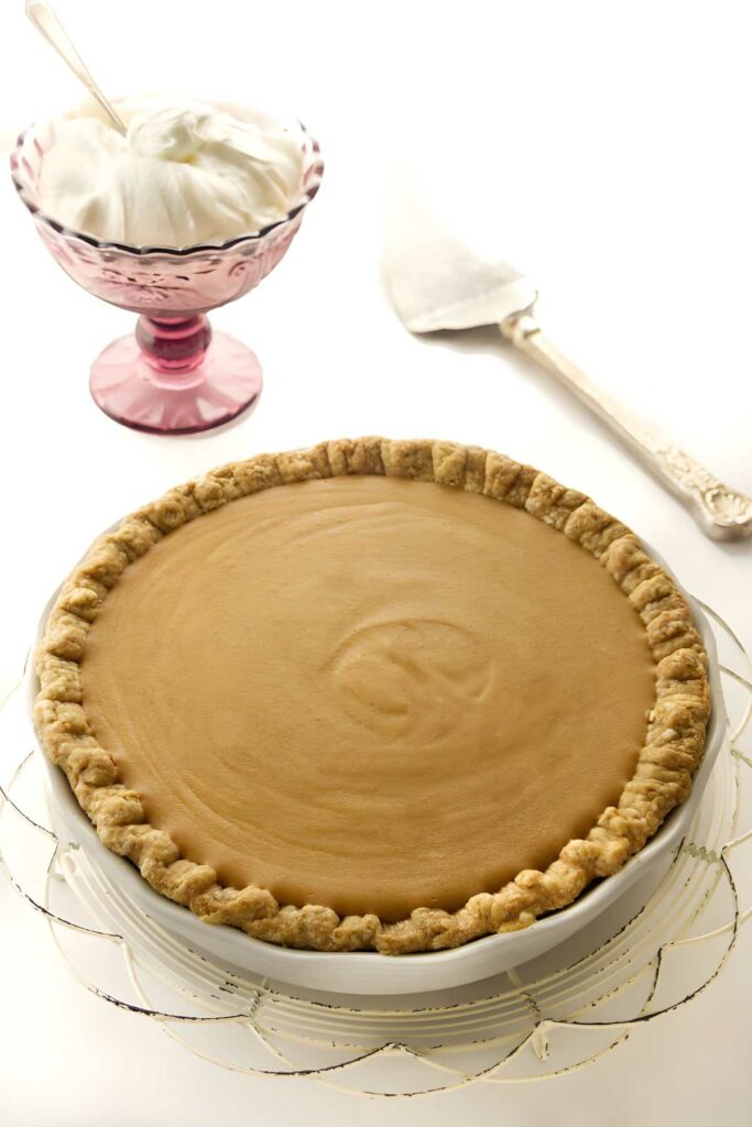 Golden-brown butterscotch pie on cake stand, vintage server and whipped cream behind.