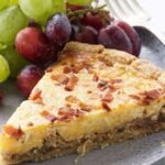 Slice of quiche with bacon on gray plate, served with grapes.