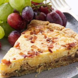 Slice of quiche with bacon on gray plate, served with grapes.