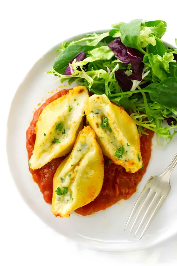 Stuffed pasta shells on tomato sauce with salad on a white plate.