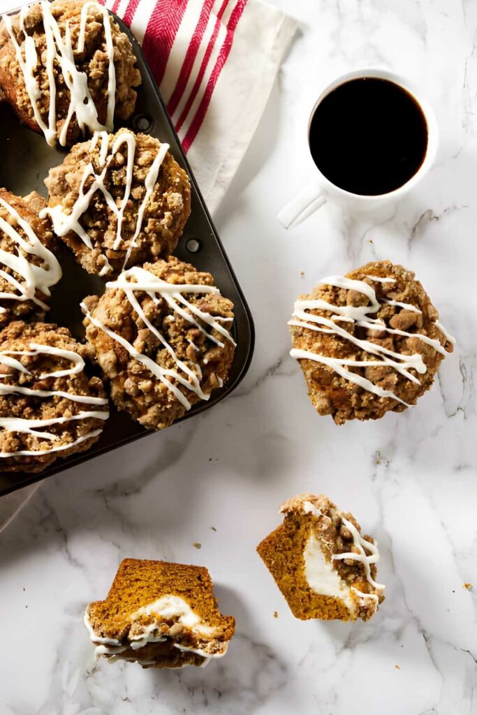 cheesecake stuffed pumpkin muffins Crumb-topped muffins with icing in tin, coffee and halved muffin on marble.