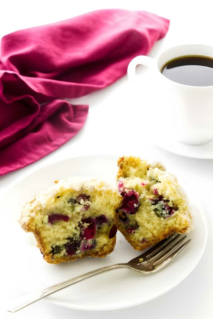 Sliced berry muffin on plate with fork, beside coffee and magenta cloth.