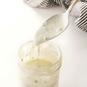 Spoon drizzling creamy herb dressing into glass jar; striped cloth background.