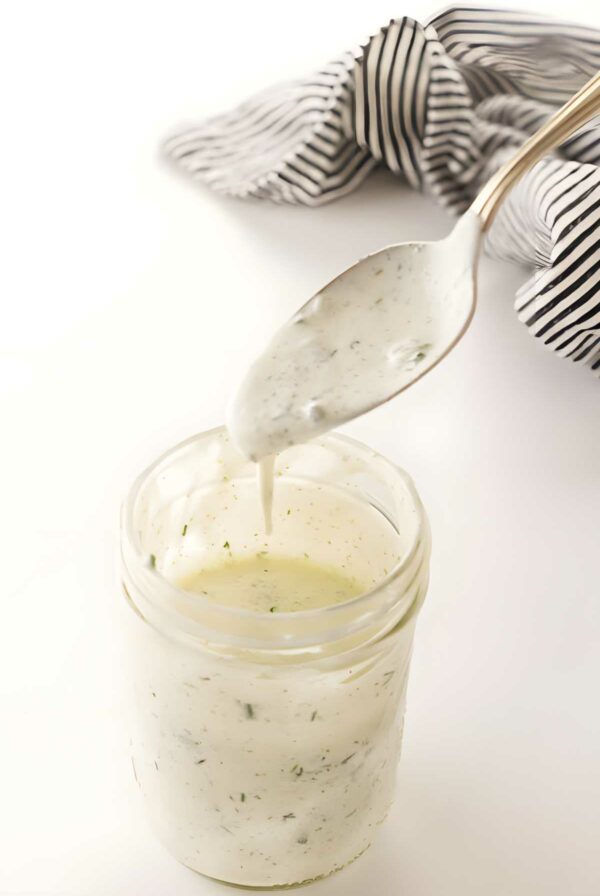 Spoon drizzling creamy ranch dressing into glass jar; striped cloth background.