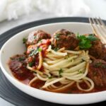 Spaghetti with tomato sauce and meatballs in a white bowl, fork beside.