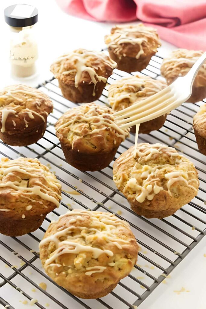 Hand drizzles icing on golden muffins cooling on a rack.