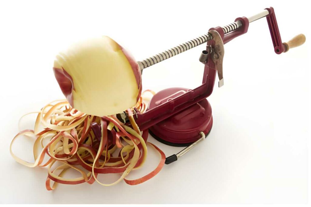 Manual apple peeler with partially peeled apple and curly peels on white background.