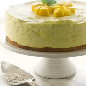 Round mango mousse cake with diced mango, mint, on a white cake stand.