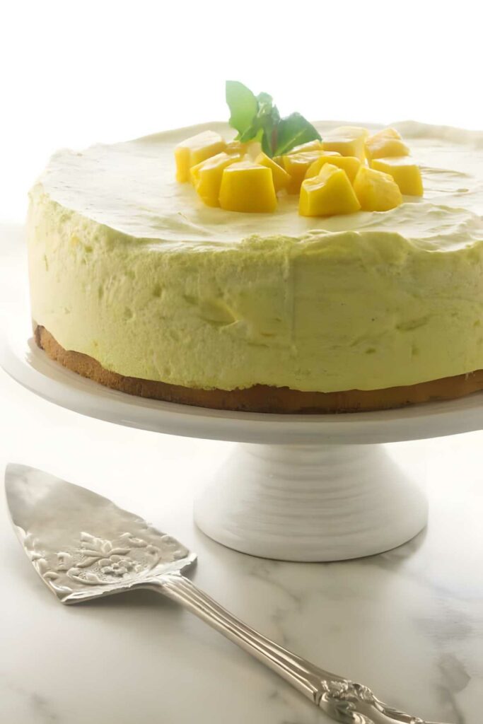Round mango mousse cake with diced mango, mint, on a white cake stand.