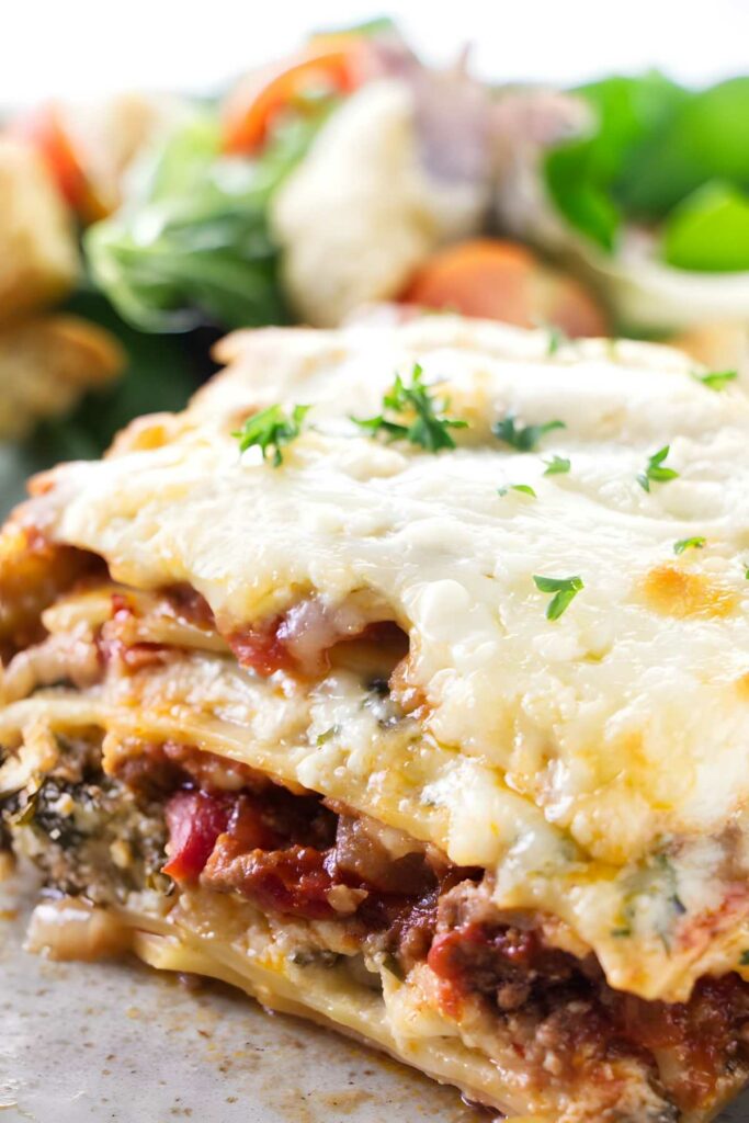 Cheesy slice of meat lasagna with layers, topped with parsley; salad in background.