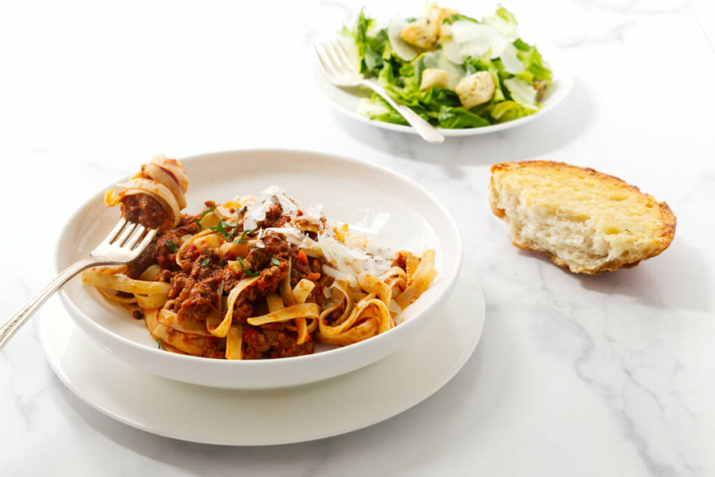 A bowl of meat based spaghetti sauce next to a salad and garlic bread.