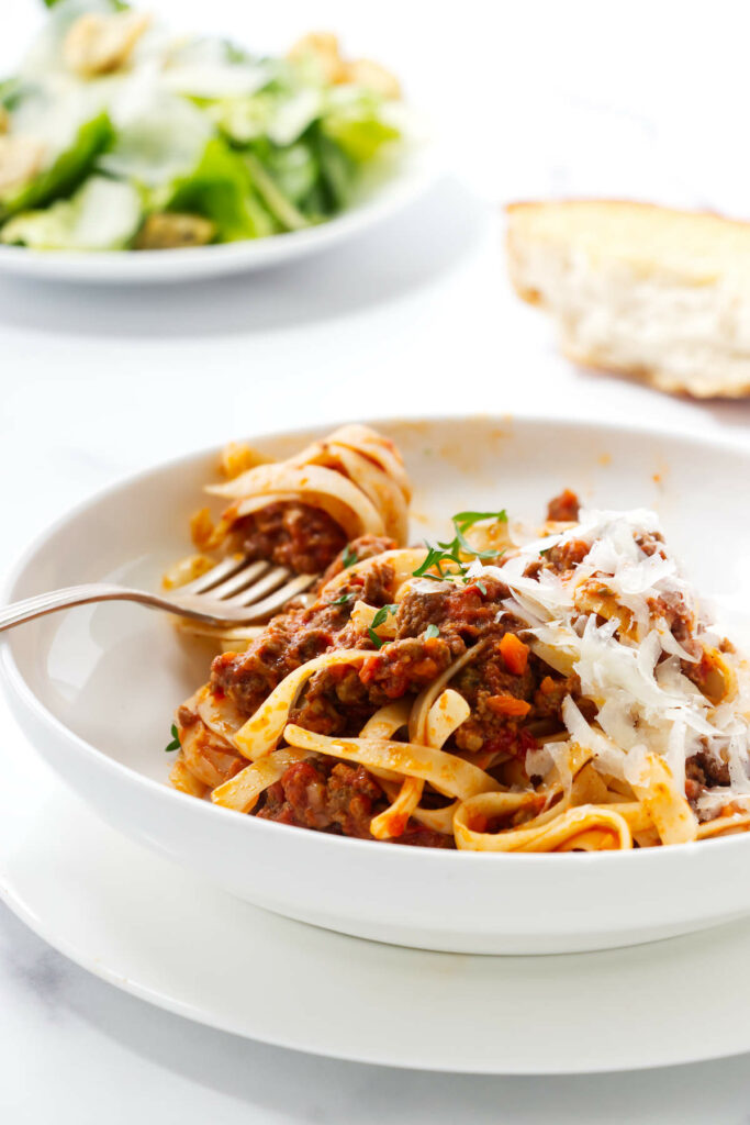 A fork in a bowl with pasta bolognese next to a salad.
