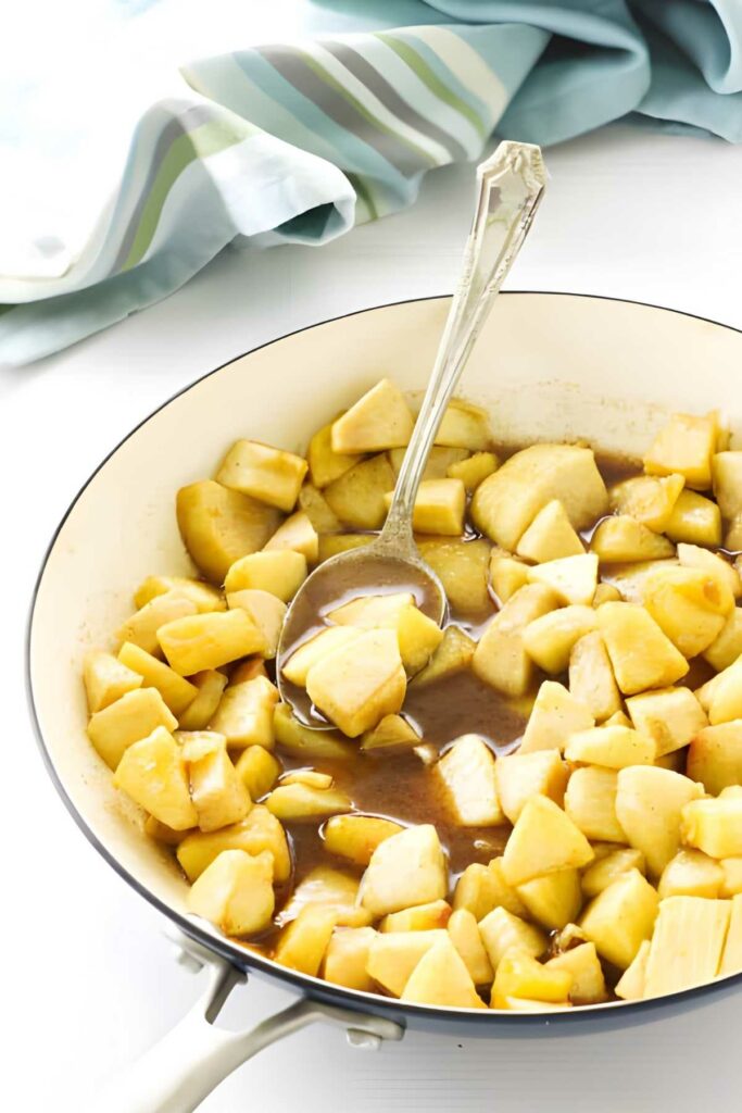 Chopped apples simmer in a white skillet with cinnamon sauce and spoon.