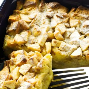 Apple french toast casserole with powdered sugar, spatula lifting a piece in black dish.