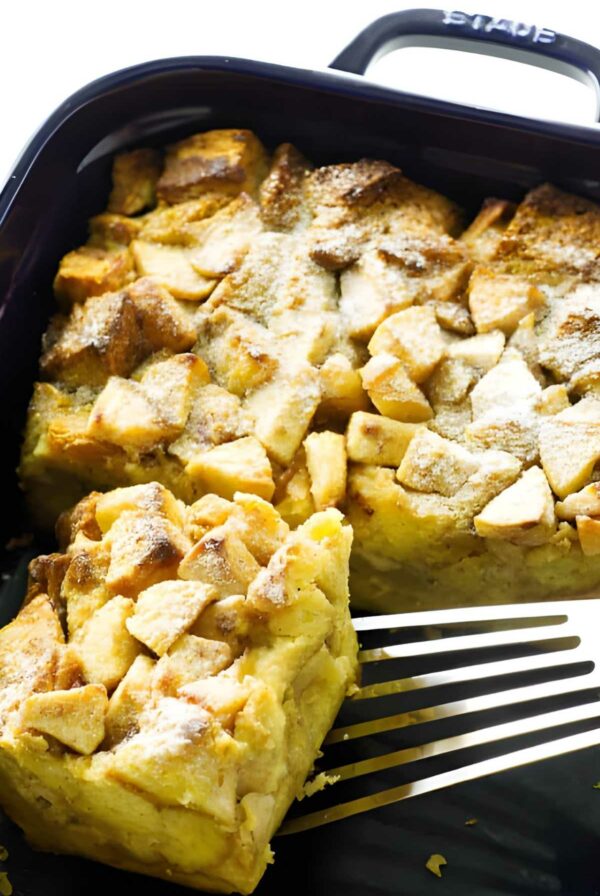 Apple french toast casserole with powdered sugar, spatula lifting a piece in black dish.