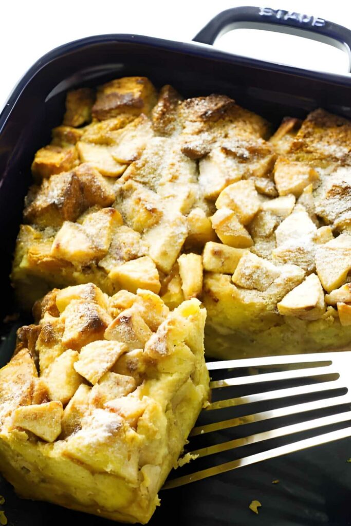 Apple french toast casserole with powdered sugar, spatula lifting a piece in black dish.