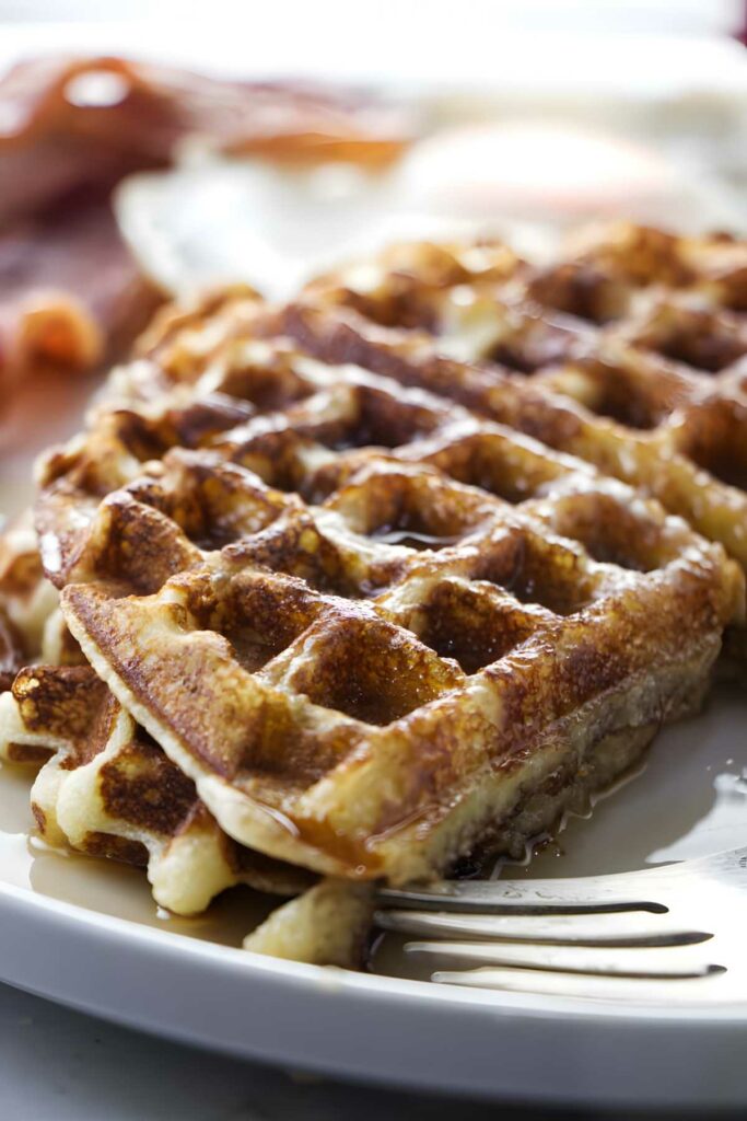 Two golden waffles with syrup on white plate, fork, blurred breakfast background.
