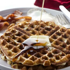 Close-up of waffles with syrup, butter, bacon, cream, and utensils.