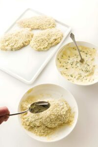 Hand breading chicken cutlets with panko; batter bowl and breaded pieces nearby.