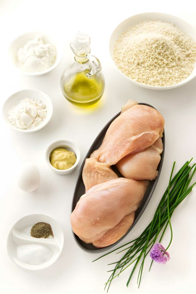 Raw chicken breasts with breading ingredients and seasonings on a white background.