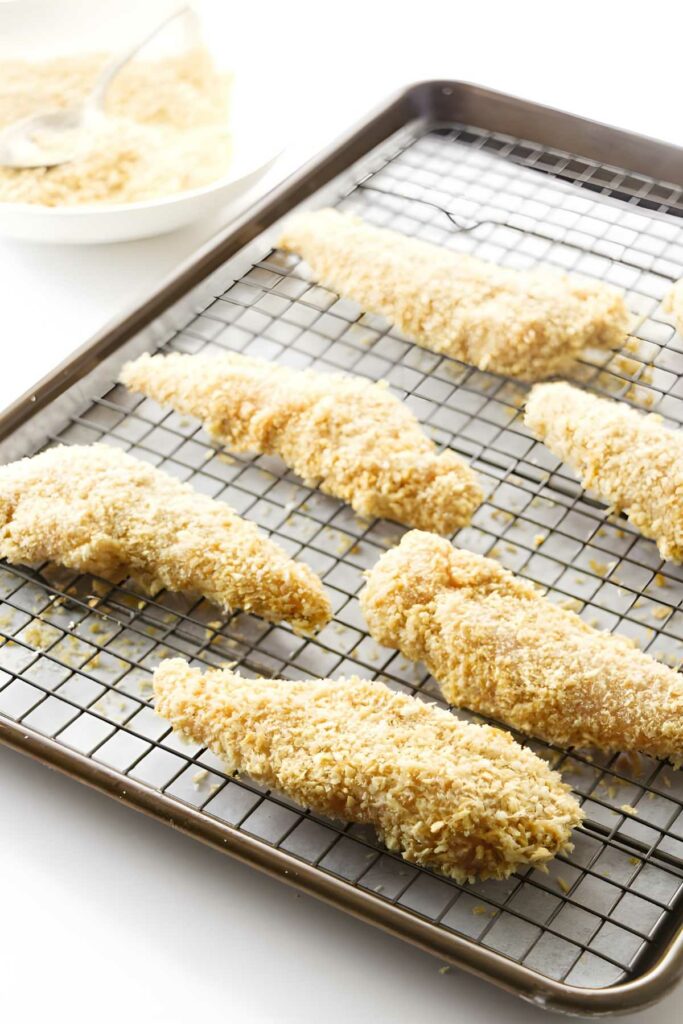 Raw breaded chicken tenders on wire rack, breadcrumbs bowl in background.