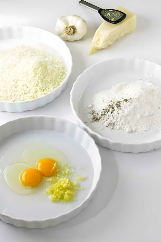 Three plates with cheese, flour, eggs, and garlic; cheese wedge and herbs nearby.