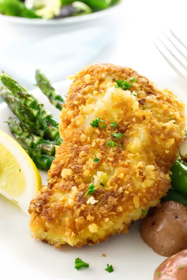 Golden-brown baked chicken cutlet with parsley, asparagus, potatoes, and lemon on plate.