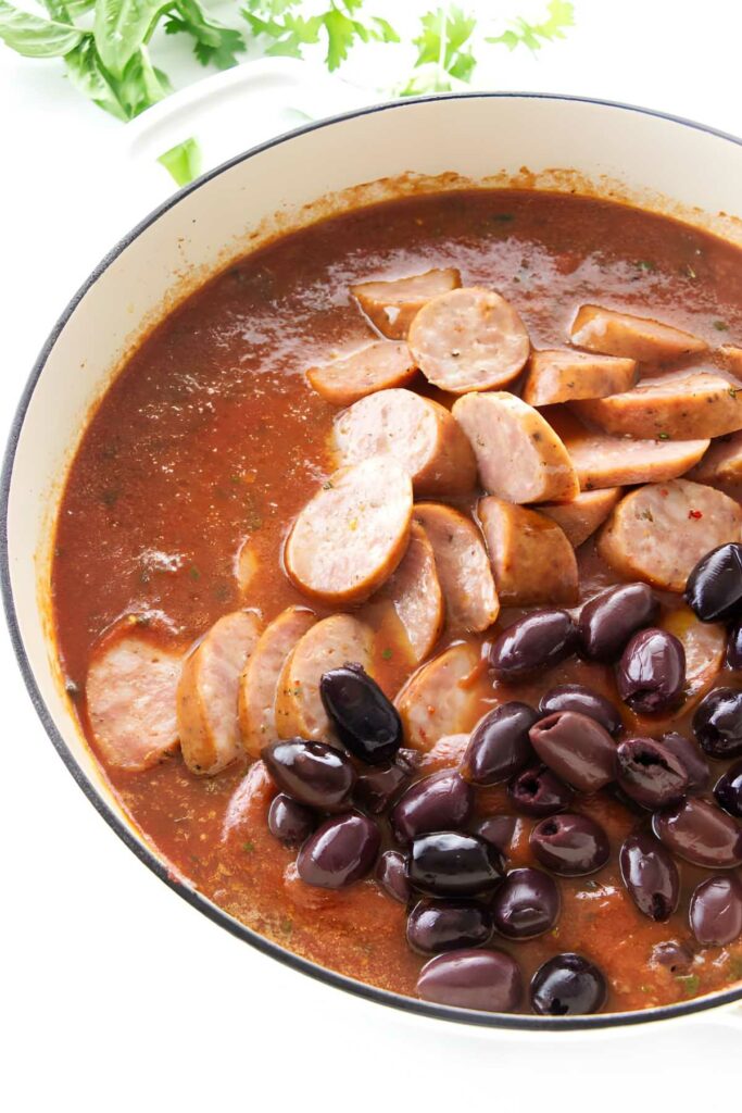 Sliced sausage and olives in tomato sauce, herbs in background.