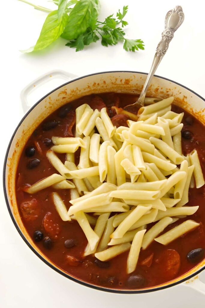 Pot of spicy tomato sauce with sausage, olives, penne pasta, and fresh herbs nearby.