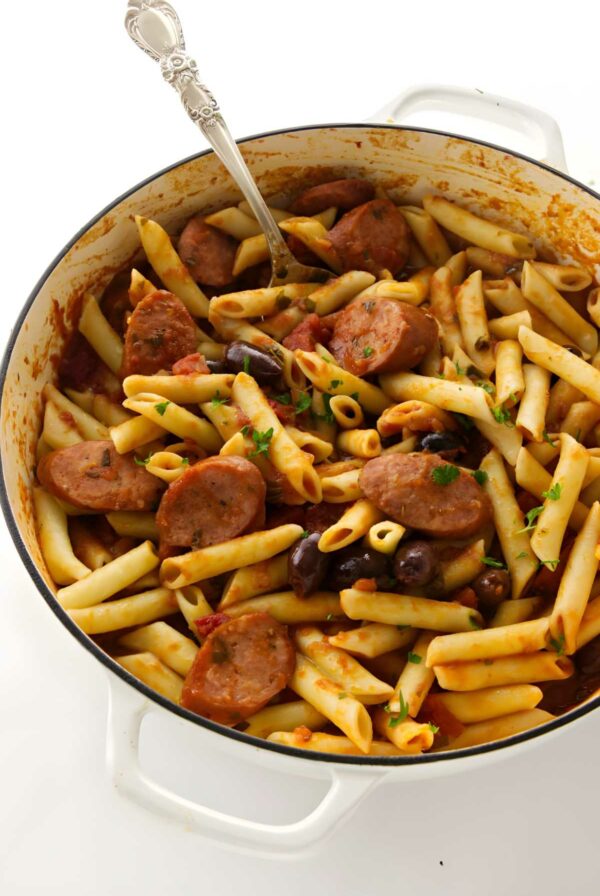 White pot with penne pasta, spicy tomato sauce, sausage, olives, tomato sauce, herbs, and spoon.