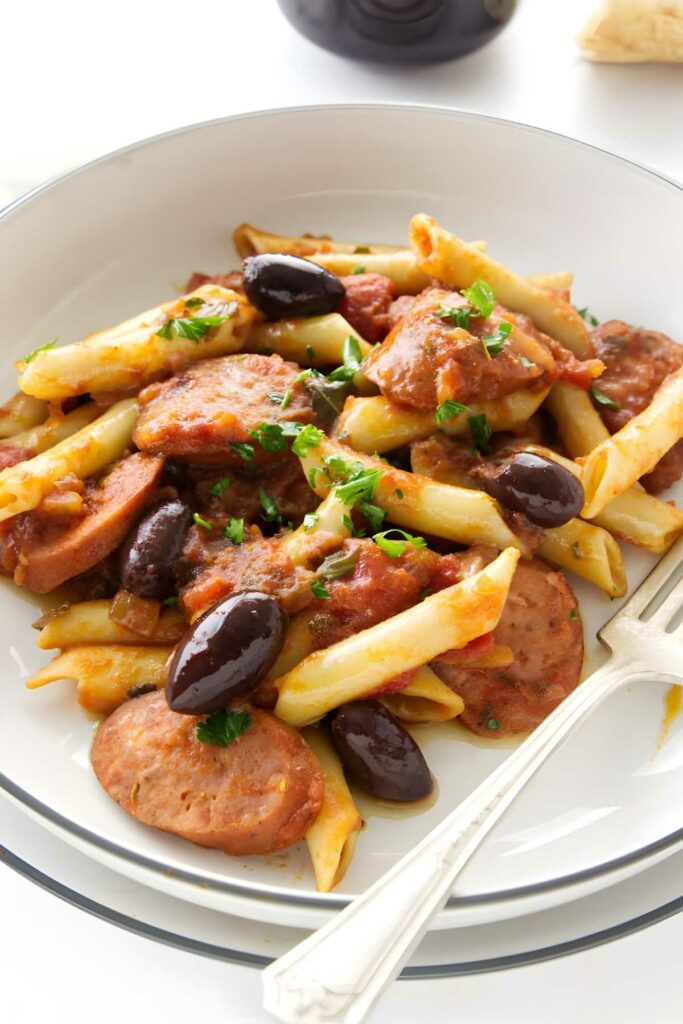 Penne pasta with sausage, olives, spicy tomato sauce, and parsley on a plate.