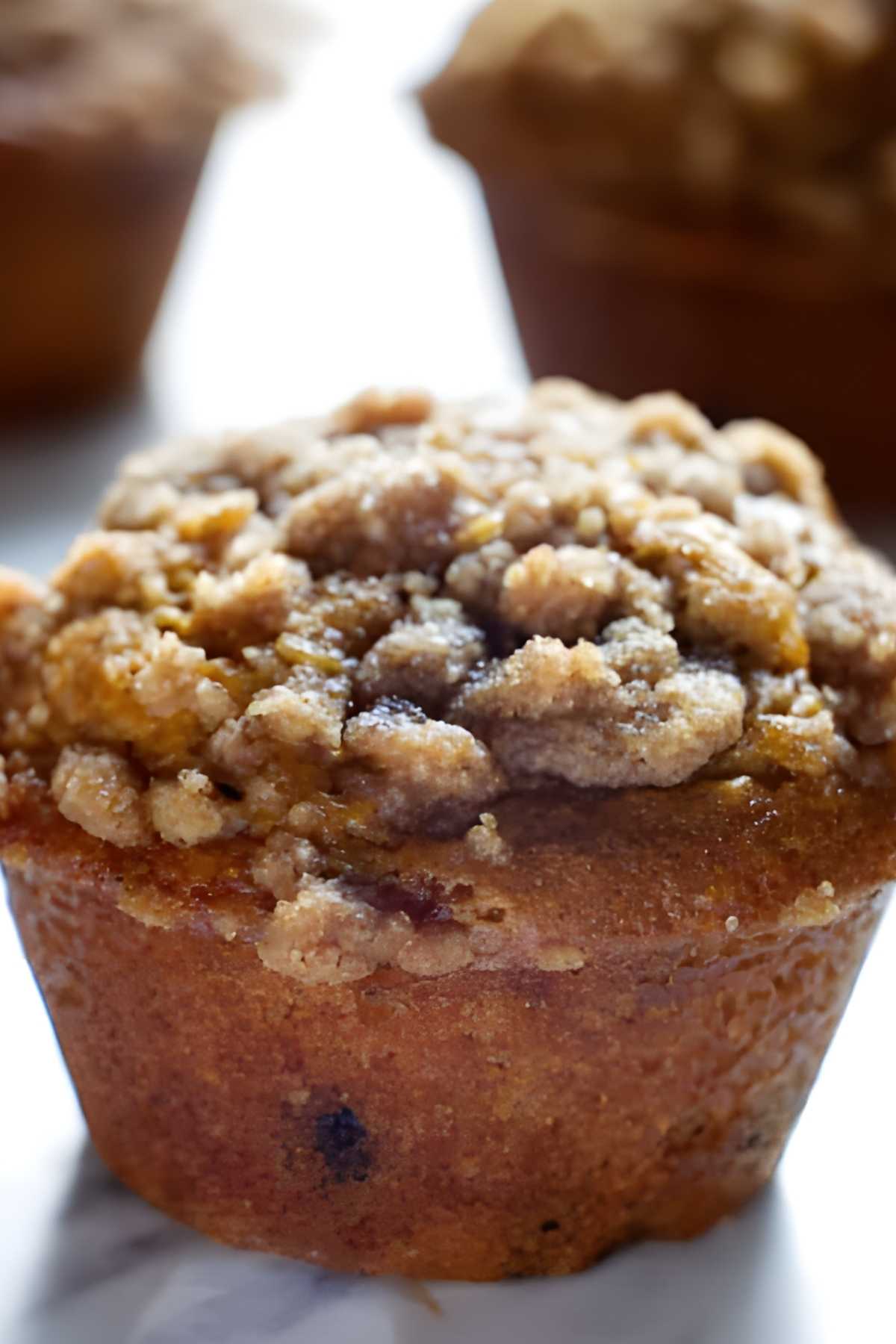 Close-up of a pumpkin muffin with crumbly streusel, two blurred in background.