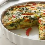Vegetable quiche with a slice missing in a white ceramic dish.