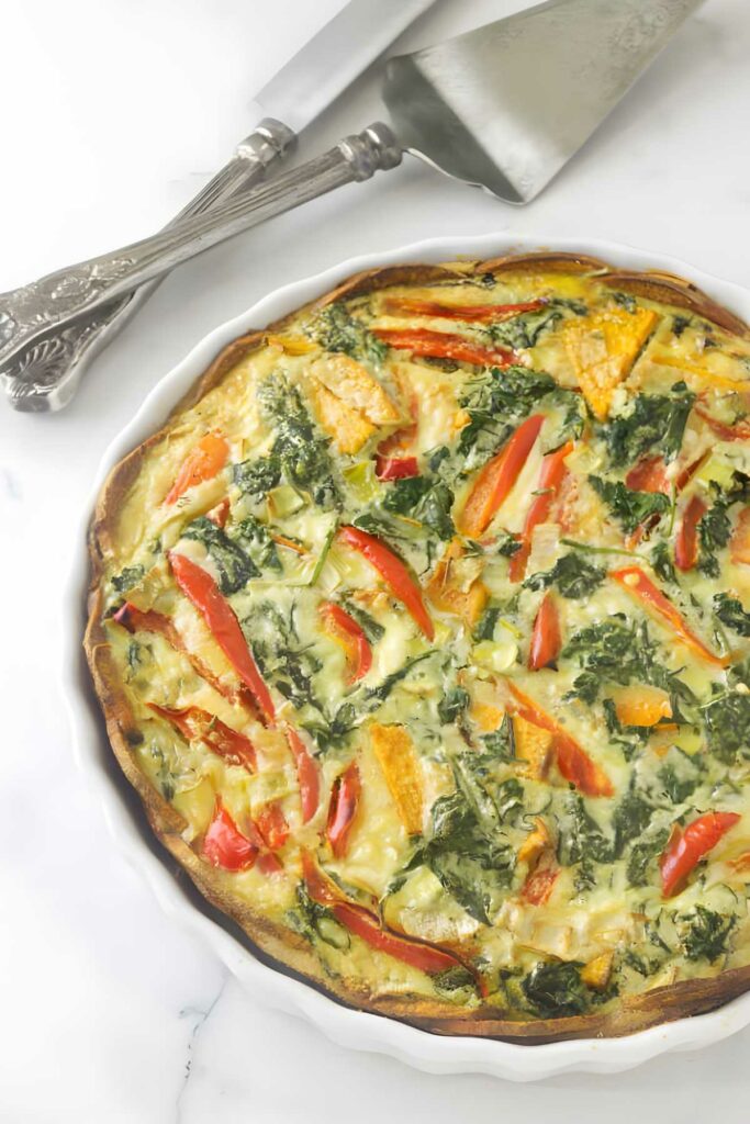 Vegetable quiche with red peppers and kale in a white dish on marble.