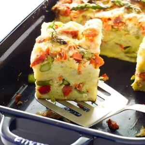 Slice of savory bread pudding with asparagus and red pepper on spatula.