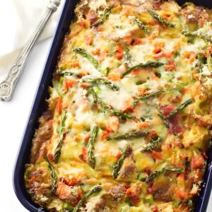 Casserole topped with cheese, asparagus, peppers, in blue dish; serving spoon nearby.