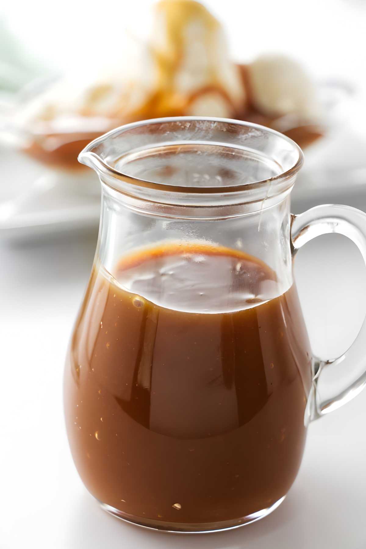 Glass pitcher of caramel sauce on white surface; blurred ice cream behind.