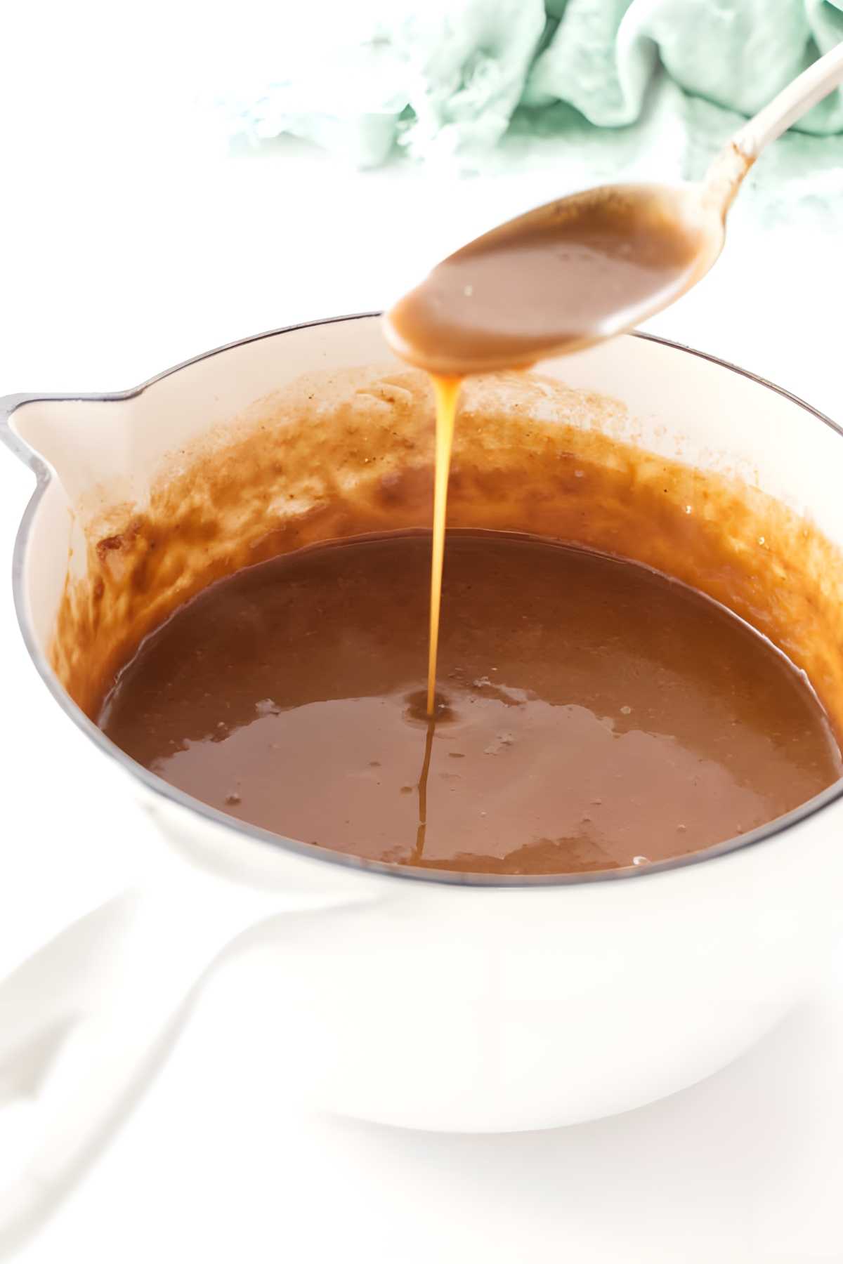 A spoon drizzles caramel sauce into a white saucepan.