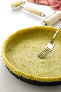 Hand docking unbaked pie crust in pan; pastry roller in background.