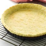 Golden baked pie crust in tart pan on cooling rack, pink cloth behind.