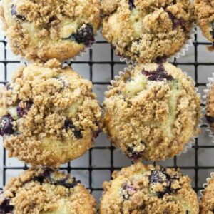 Nine golden blueberry muffins with crumb topping cooling on a black wire rack.