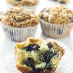 Halved blueberry muffin with juicy filling, whole muffins in background on white surface.