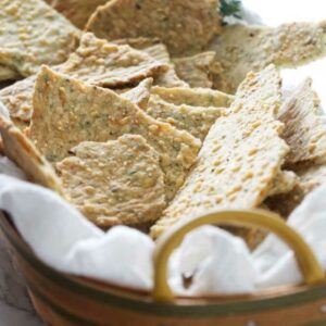 Basket with homemade seeded crackers on white cloth, herbs in background.