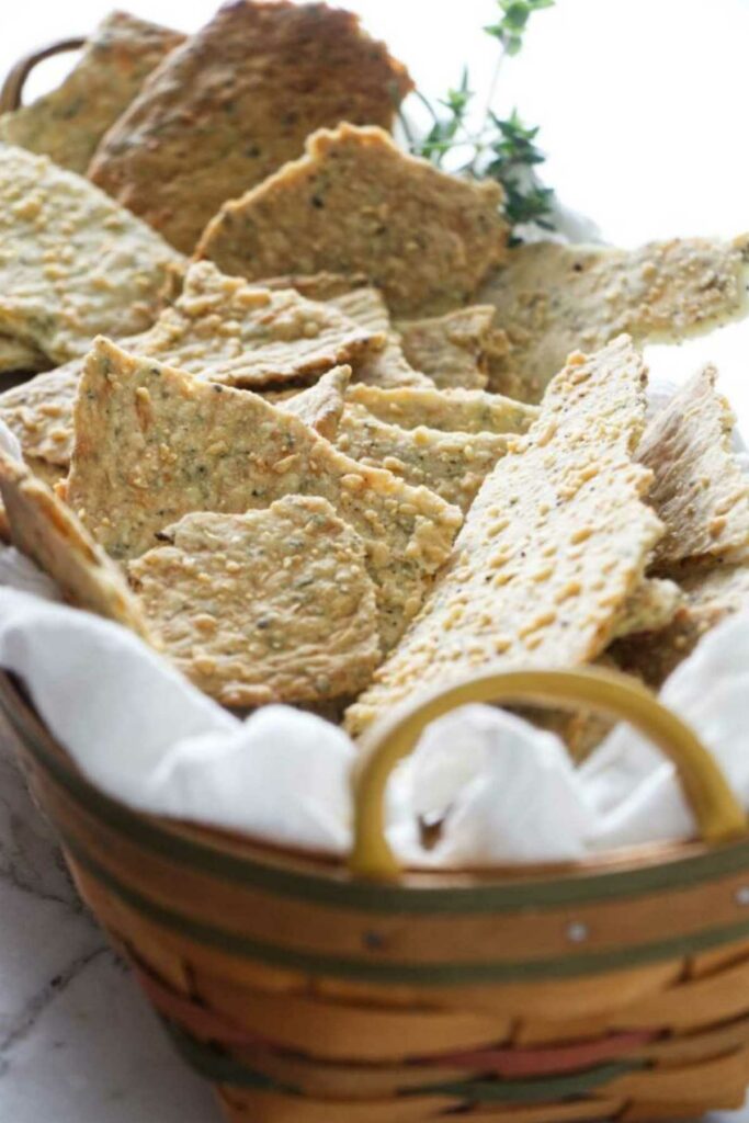 Basket with homemade seeded crackers on white cloth, herbs in background.