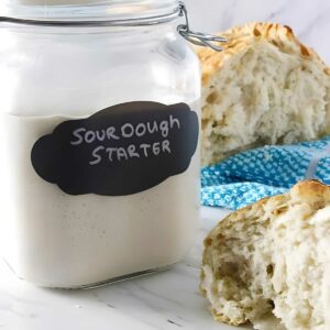 Glass jar of sourdough starter beside sliced bread and blue cloth.
